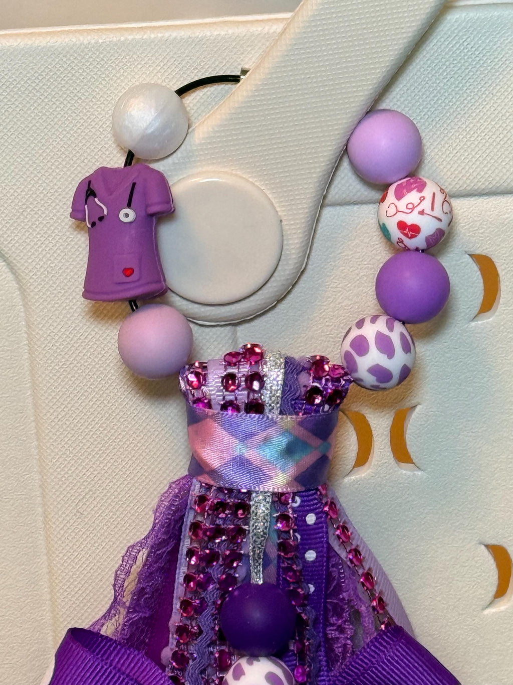 Close-up of purple Nurse Life bag tassel top showing wire loop clip with purple scrubs charm with stethoscope, medical heart-print bead, purple leopard print bead, and solid lavender and purple silicone beads over white Hurley tote bag handle, with purple rhinestone-trimmed ribbon and purple lace tassel top visible below