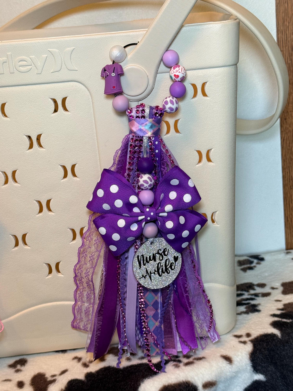 Purple Nurse Life silicone beaded bag tassel clipped to white Hurley tote bag handle on cowhide rug, featuring purple silicone bead strand with purple scrubs charm, medical heart-print bead, leopard print bead, and solid lavender and purple beads, purple polka-dot grosgrain bow, cascading purple multi-ribbon tassel with rhinestone trim, and silver glitter Nurse Life pendant with EKG heartbeat detail