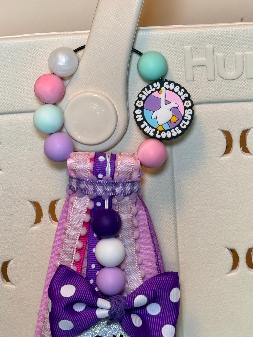 Close-up of Silly Goose On The Loose Club silicone bead charm and pastel pink, mint, lavender, and purple silicone beads on a bag tassel clipped onto a white tote bag handle

