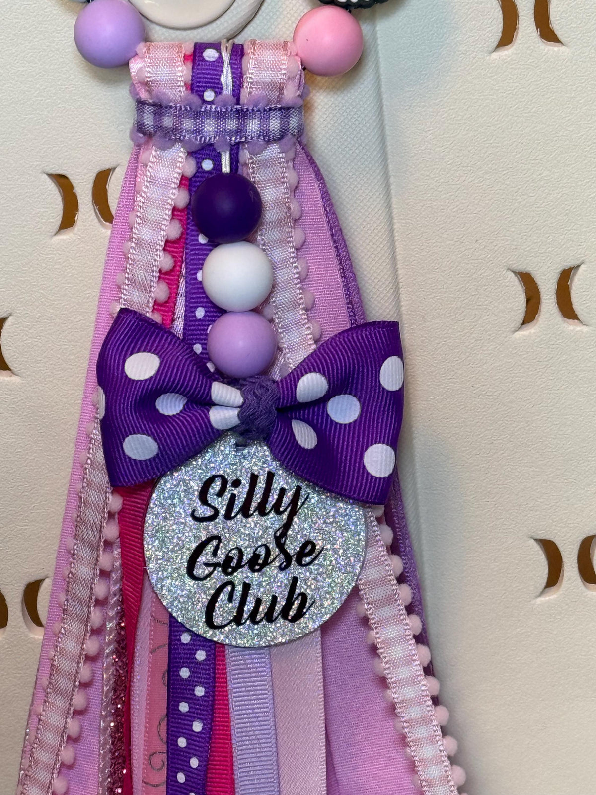Close-up of glitter Silly Goose Club charm disc with purple polka dot bow and layered pink, purple, and lavender ribbon tassel on a silicone beaded bag tassel
