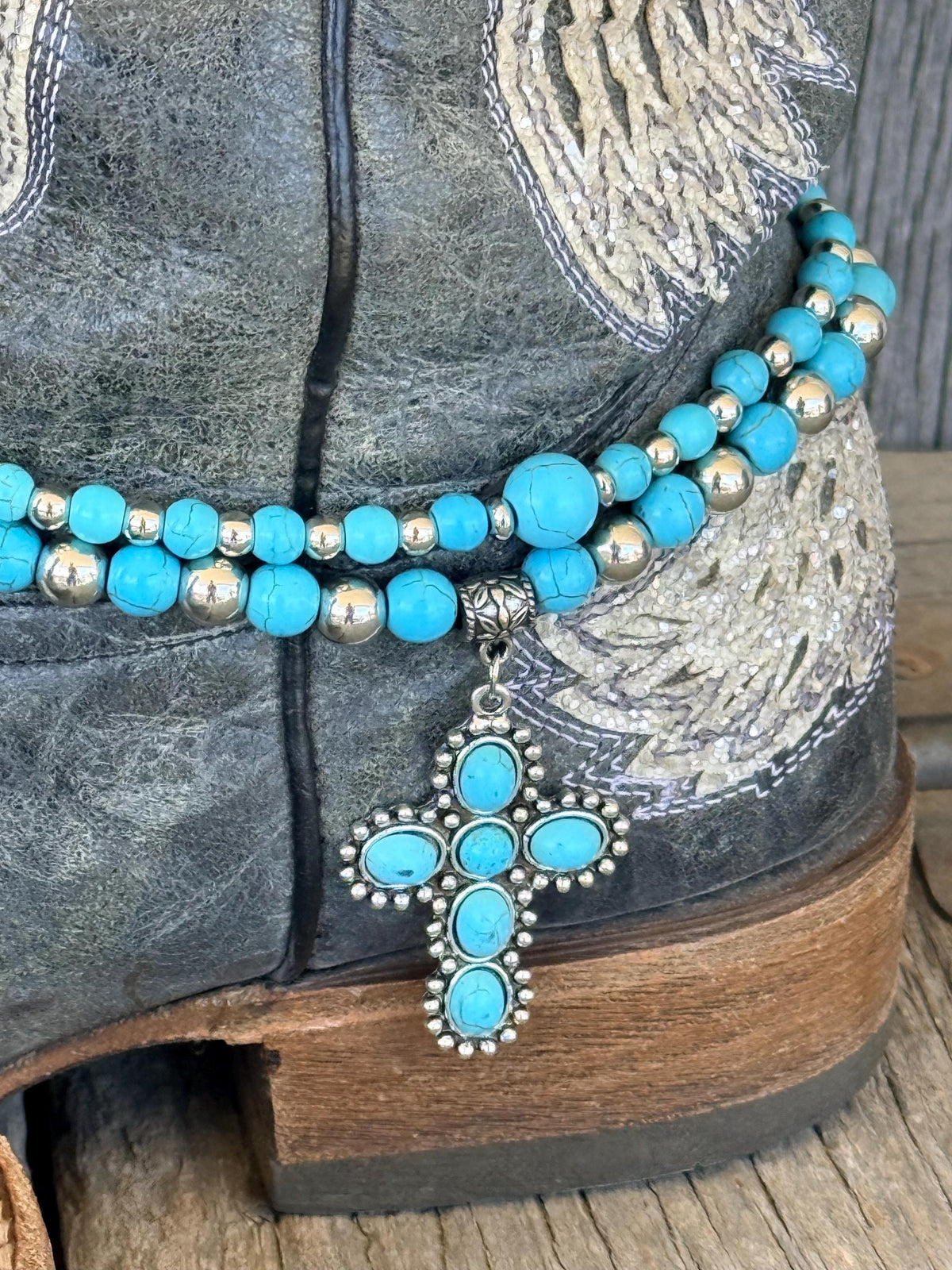 Side angle close-up of turquoise and silver beaded boot bracelet with large turquoise stone cross charm on black angel wing cowboy boot