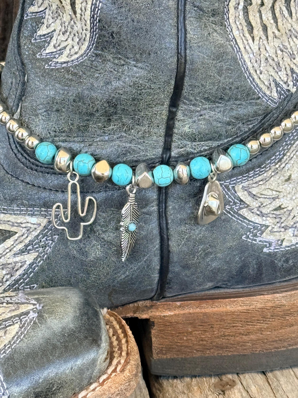 Angled front view of black distressed angel wing cowboy boots on wood surface with turquoise stone and silver bead boot bracelet showing silver cactus, feather, and cowboy hat charms