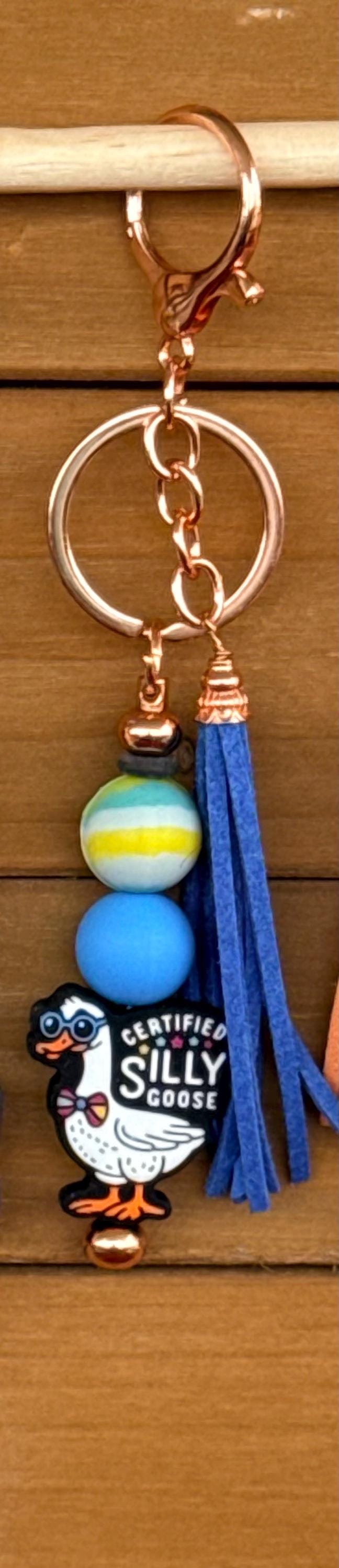 A close-up of a keychain with a copper-tone clasp and split ring, featuring a blue tassel, a blue and yellow bead, a small blue ball, and a Goose charm labeled 'Certified Silly Goose' against a wooden background.