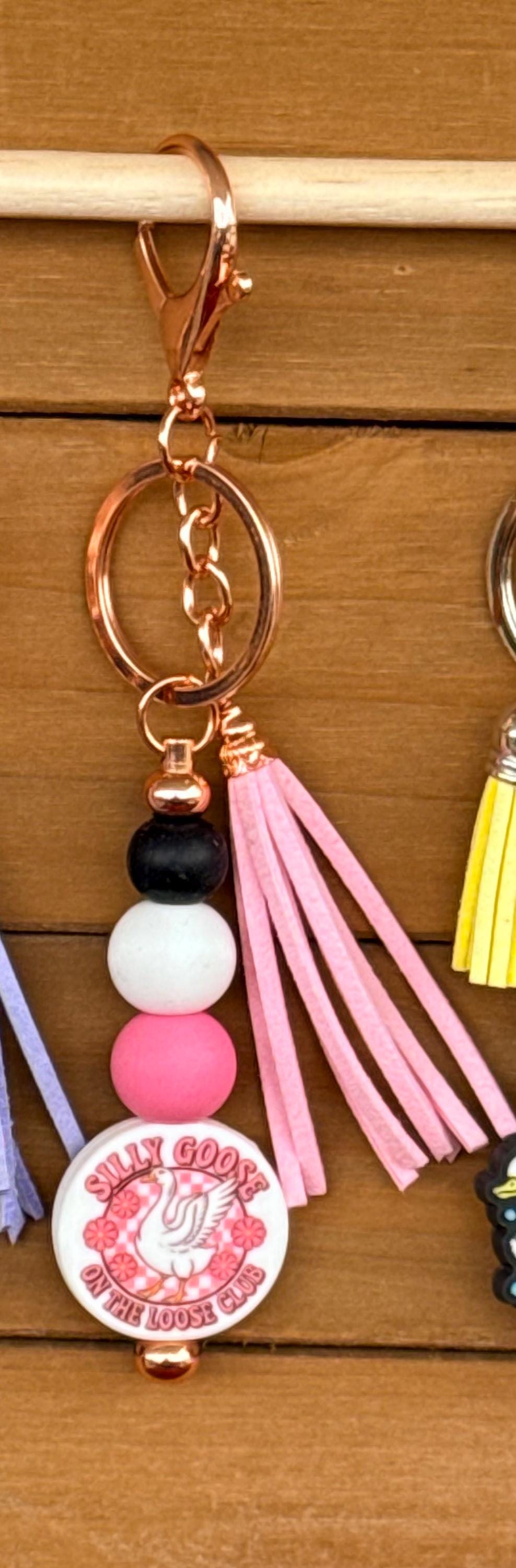 A close-up of a keychain with a rose gold clasp and key ring, featuring pink and white beads, a pink faux-leather tassel, and a round white charm with a pink graphic that reads Silly Goose On The Loose Club