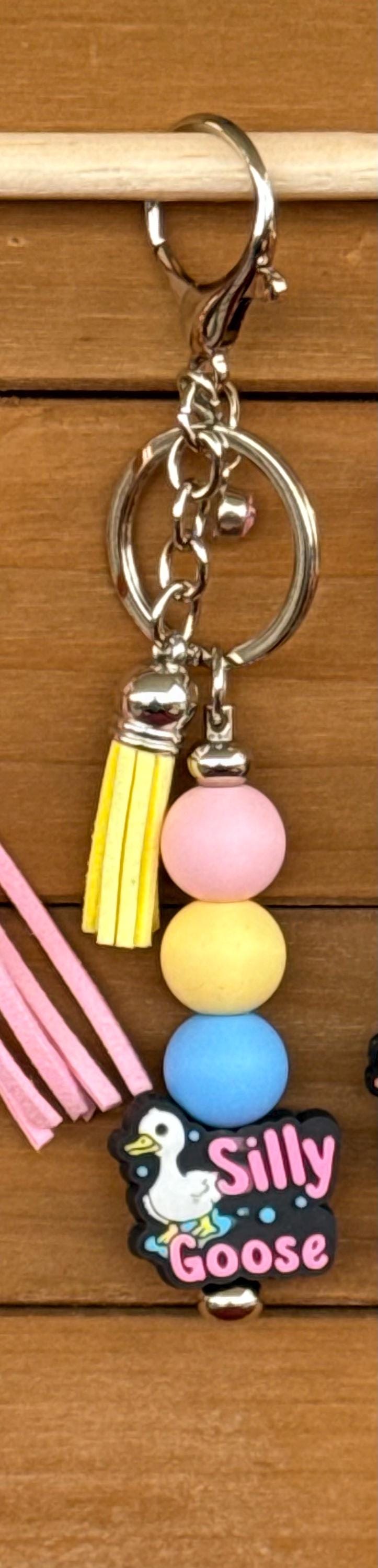 A novelty keychain featuring a silver clasp, a yellow tassel, three pastel-colored beads (pink, yellow, blue), and a small black charm shaped like a goose with the text 'Silly Goose'.