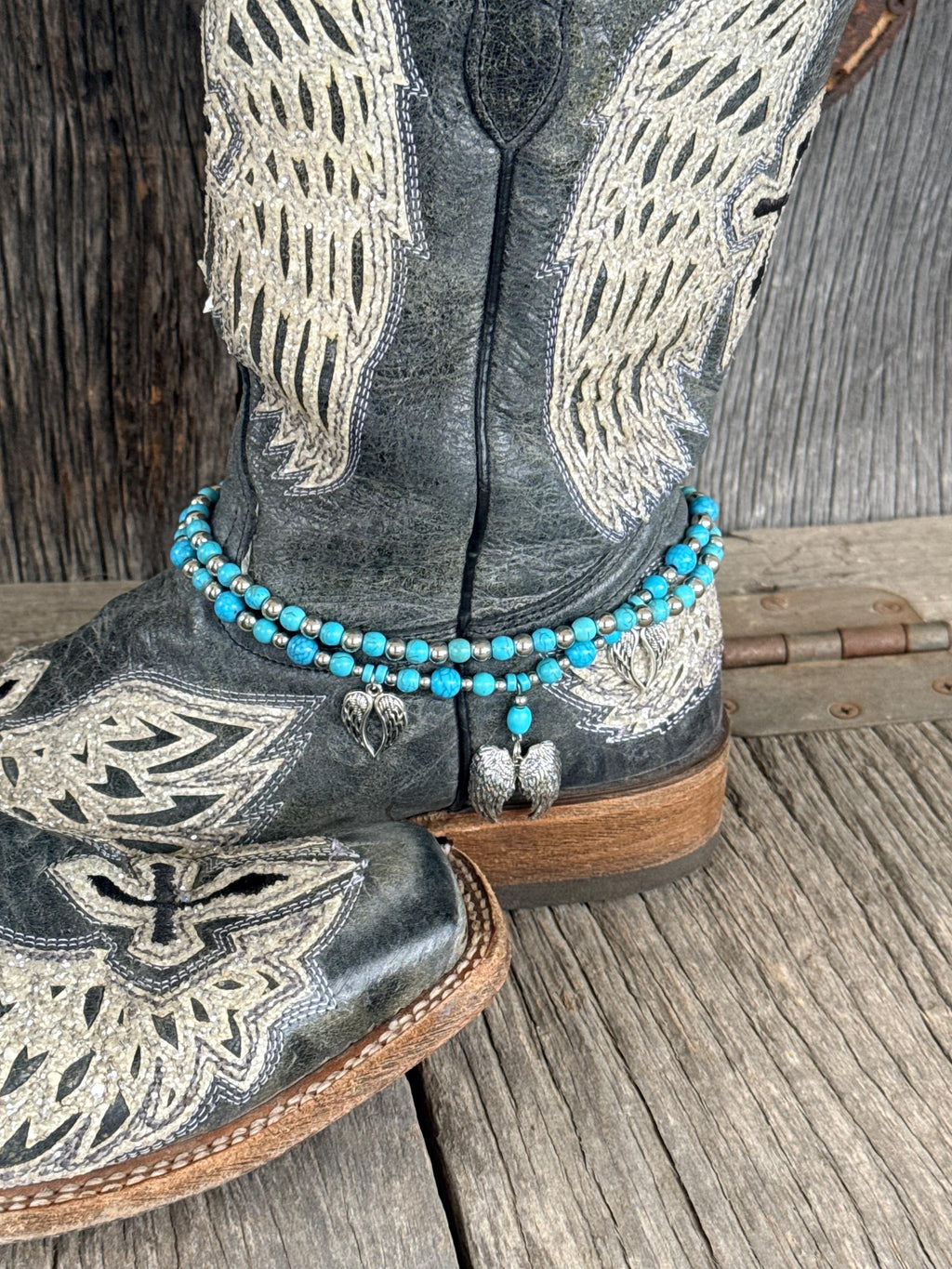 A close-up of a western boot with turquoise beaded ankle bracelets and silver angel wing charms, resting on a rustic wooden surface.