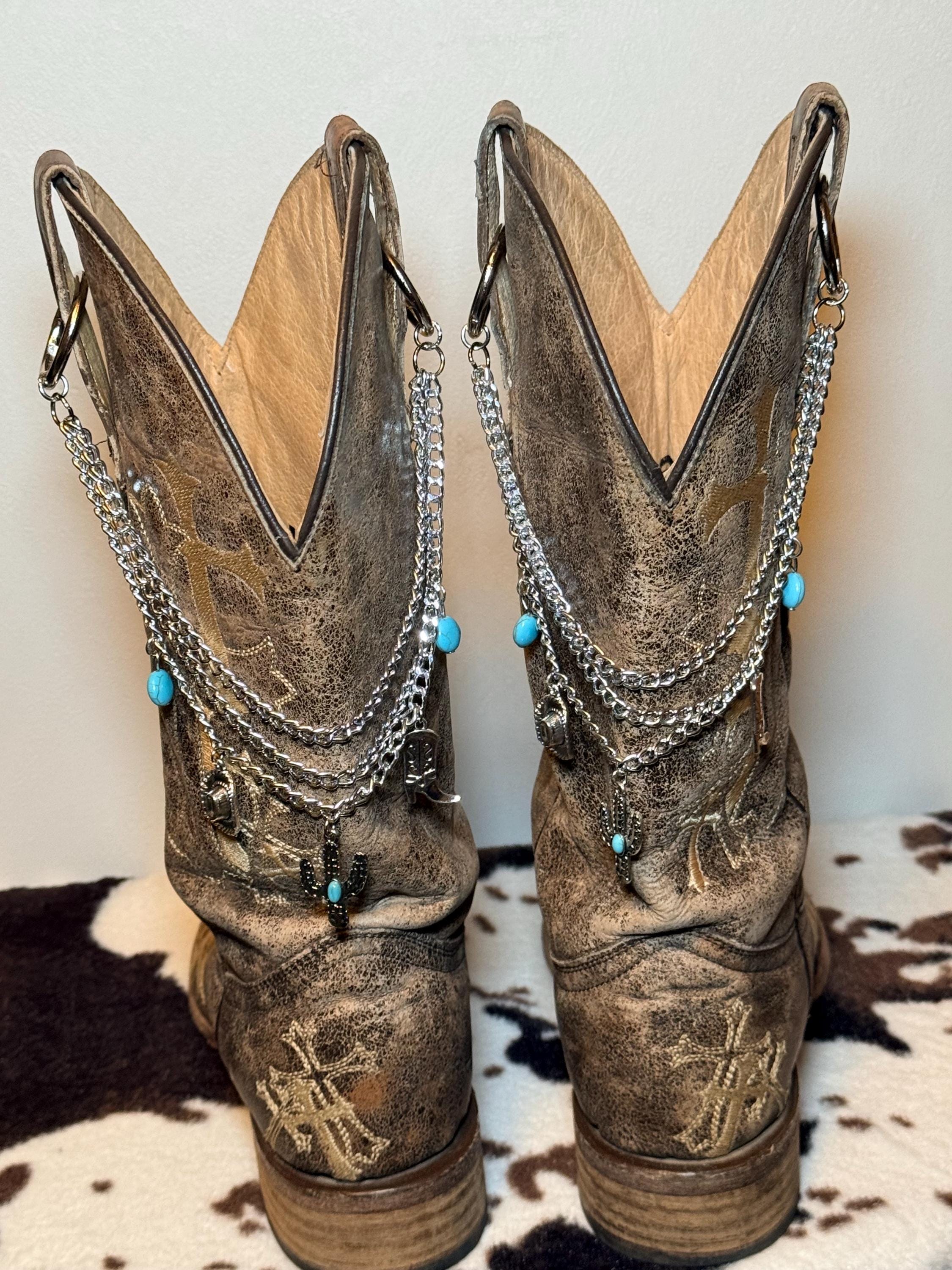 Front view of pair of tan distressed cross-embroidered cowboy boots on cowhide rug wearing three-strand silver chain lei boot bracelets with turquoise oval bead accents and silver Western charms including cactus, cowboy hat, and cowboy boot