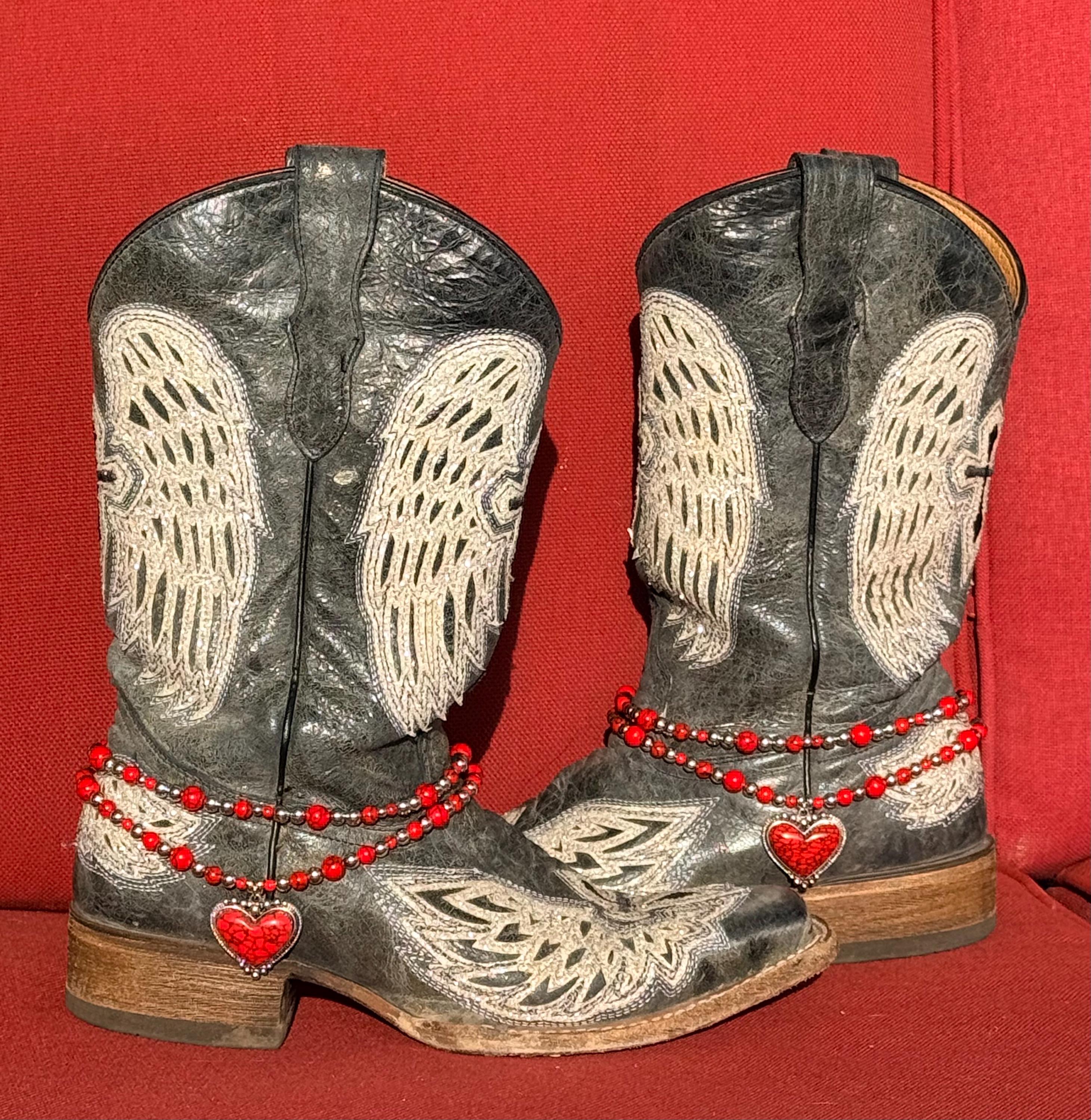 Front view of pair of black distressed angel wing laser-cut cowboy boots against red background wearing two-strand red round bead and silver ball spacer boot bracelet chains with large red crackle stone heart pendant in silver bezel frame, Western boot bling set of 2