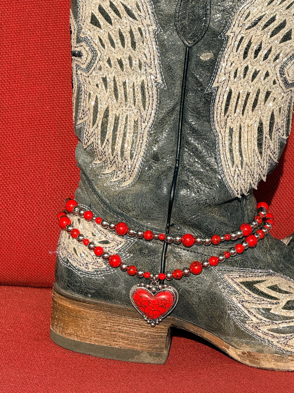Close-up of black distressed angel wing laser-cut cowboy boot ankle with two-strand red round bead and silver ball spacer boot bracelet chain featuring large red crackle stone heart pendant set in silver bezel frame