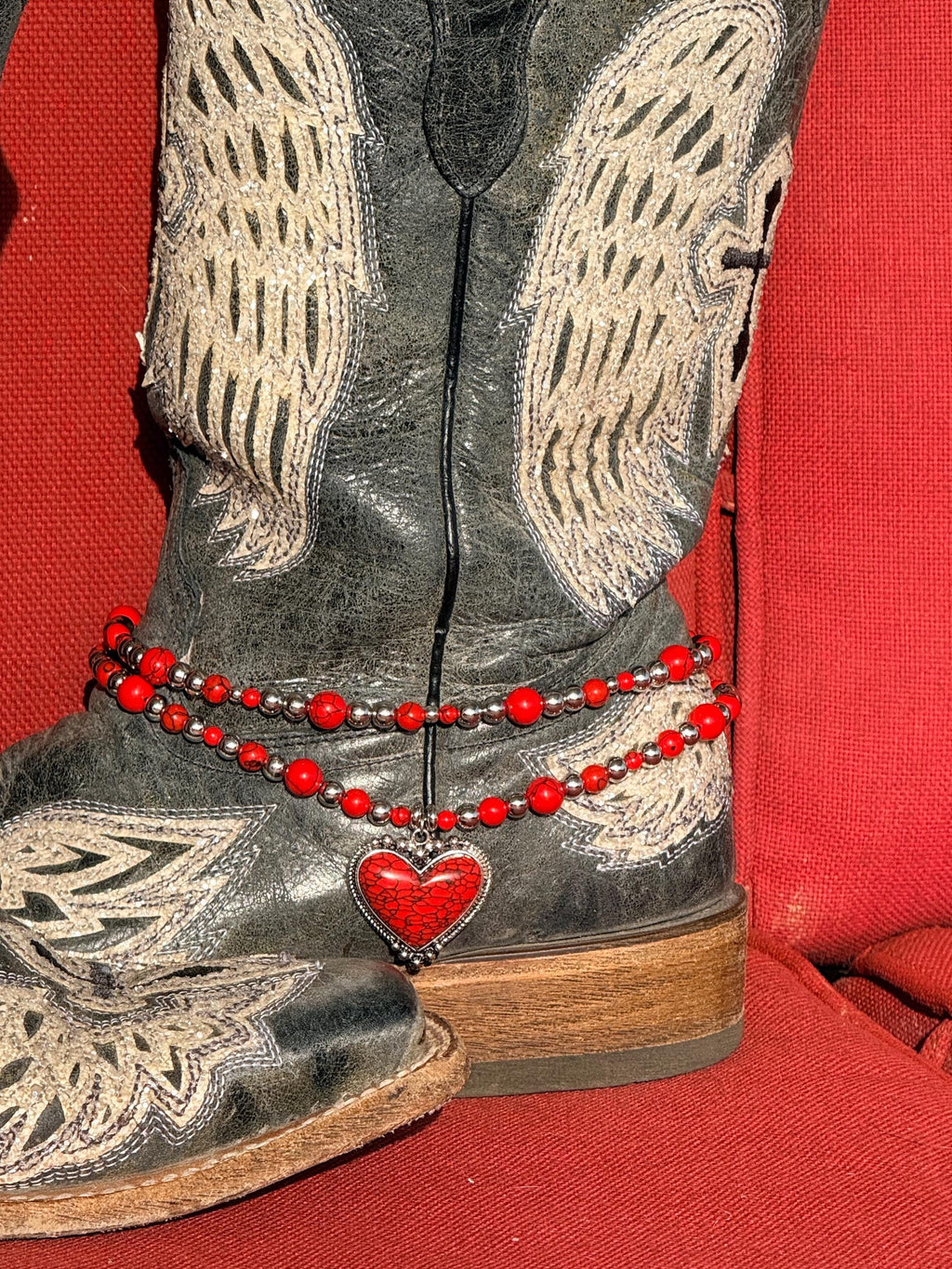 Front view of black distressed angel wing and cross-embroidered cowboy boot with two-strand red round bead and silver ball spacer boot bracelet chain and large red crackle stone heart pendant in silver bezel frame, Western Valentine's boot jewelry