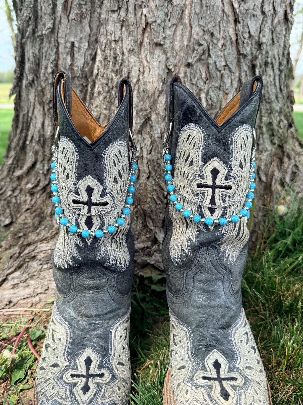 Top-down front view of pair of black cross and angel wing embroidered cowboy boots on grass against tree wearing turquoise round bead and silver ball spacer lei boot bracelets clipped through boot pull loop rings, Western boho boot jewelry set of 2