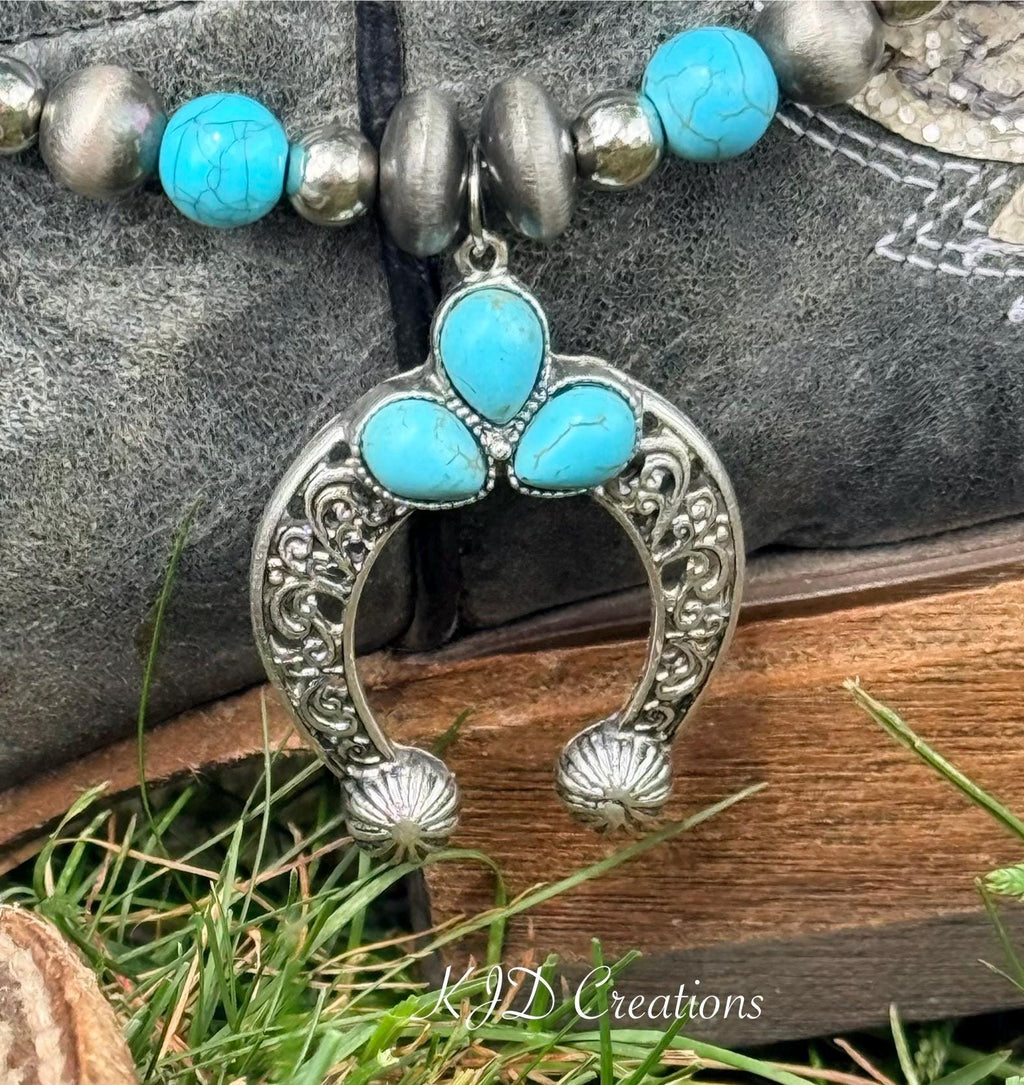 Macro close-up of silver filigree squash blossom naja pendant with three turquoise teardrop stones, horseshoe-shaped silver frame with scroll filigree and shell-tip ends, on two-strand turquoise and silver bead boot bracelet