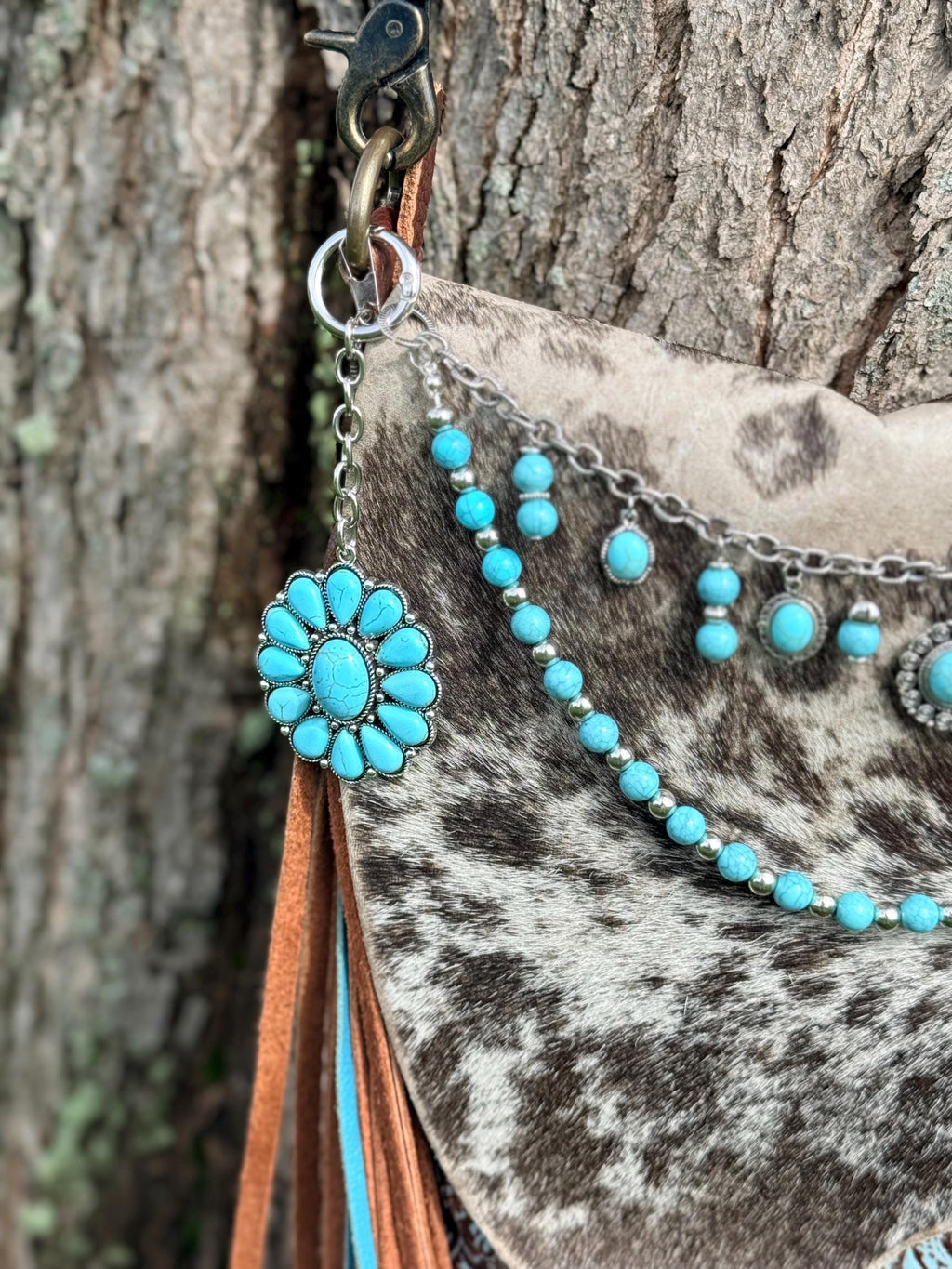 Close-up of left side hardware of brown and black cowhide crossbody bag showing large turquoise flower concho with oval center turquoise stone surrounded by teardrop turquoise stones in silver bezel setting hanging from silver ring clip, with turquoise round bead strand with silver rondelle spacers and silver cable chain with dangling turquoise cabochon cluster charms visible across bag front, Western boho purse chain detail