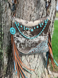 Front view of brown and black cowhide crossbody bag with tan leather fringe against tree trunk, featuring three-strand Western boho turquoise and silver purse chain with silver cable chain with nine dangling turquoise cabochon cluster charms and crystal rhinestone concho pendant at center, turquoise round bead strand with silver rondelle spacers, and large turquoise flower concho hanging from left side hardware, 9.75-inch Western handbag accessory