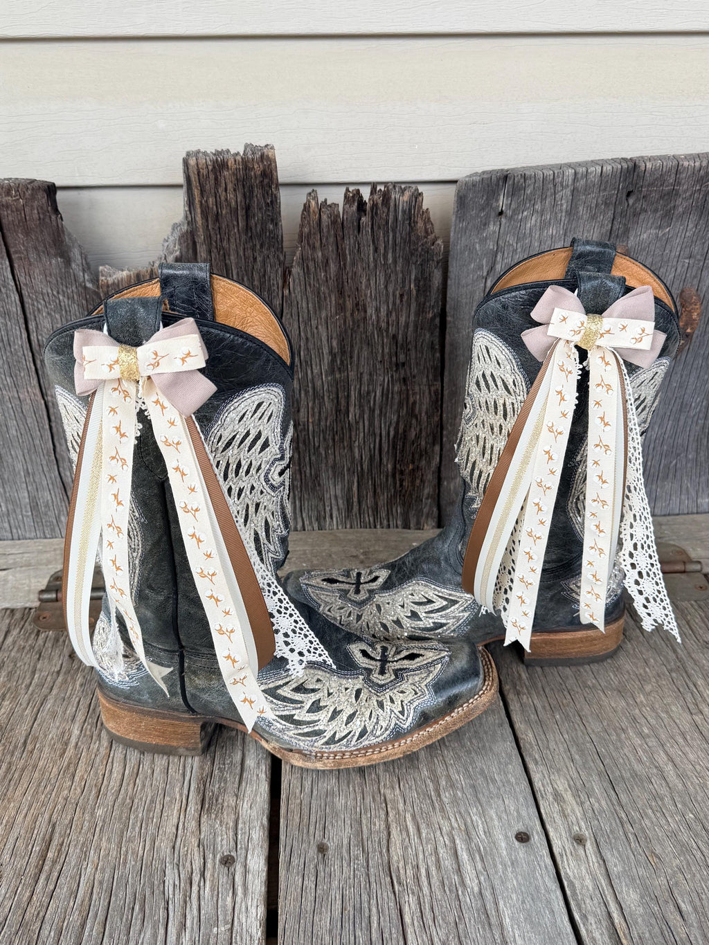 A pair of western cowboy boots with dark gray and black leather, featuring white embroidered patterns, tan trim, and long cream ribbons with gold lettering tied in bows on the shafts, displayed on weathered wooden planks.