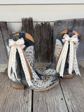 A pair of western cowboy boots with dark gray and black leather, featuring white embroidered patterns, tan trim, and long cream ribbons with gold lettering tied in bows on the shafts, displayed on weathered wooden planks.