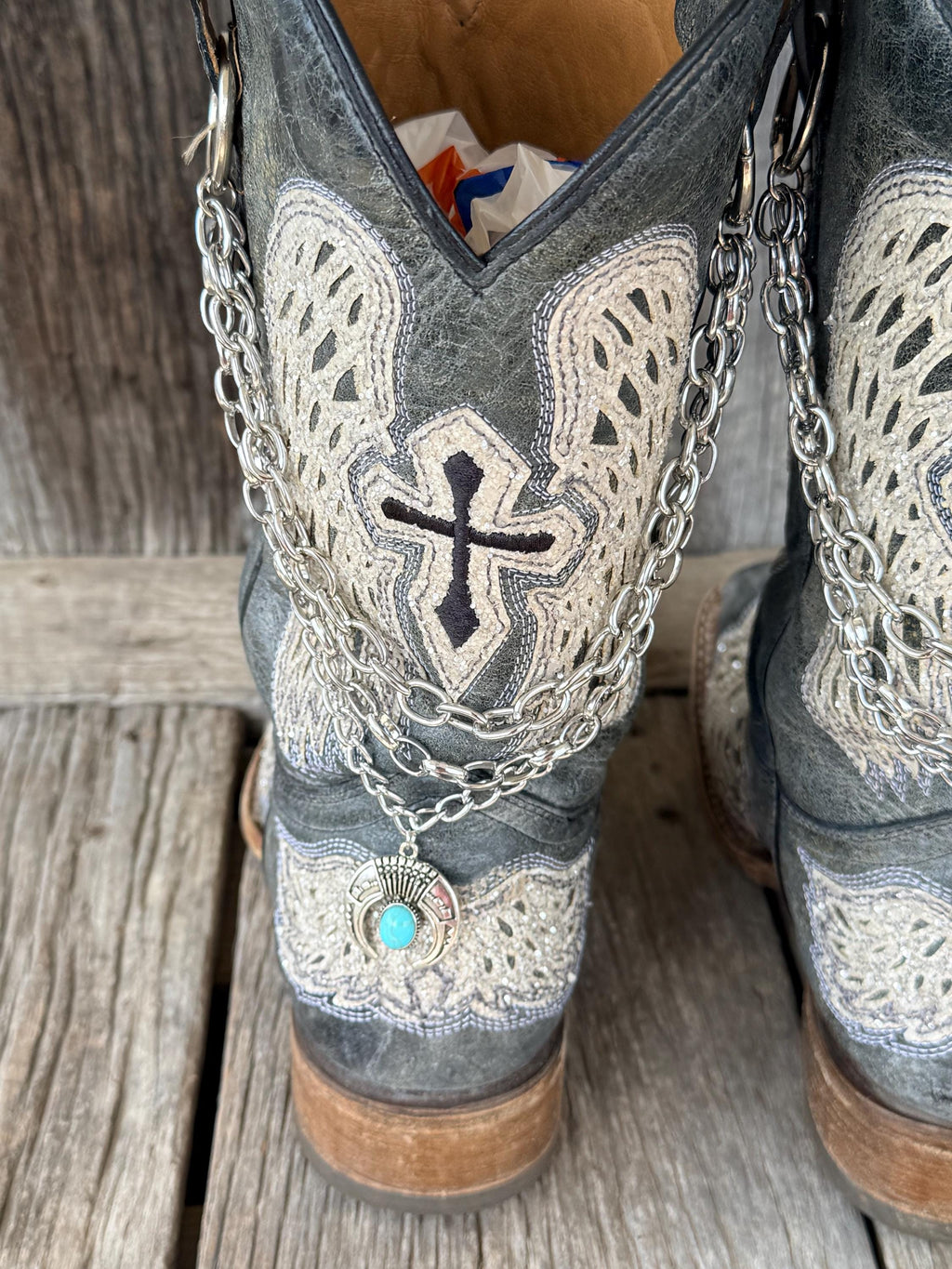 Angled front view of distressed gray cross and wing cowboy boots with layered silver chain lei boot bracelets and turquoise squash blossom charms, set of 2