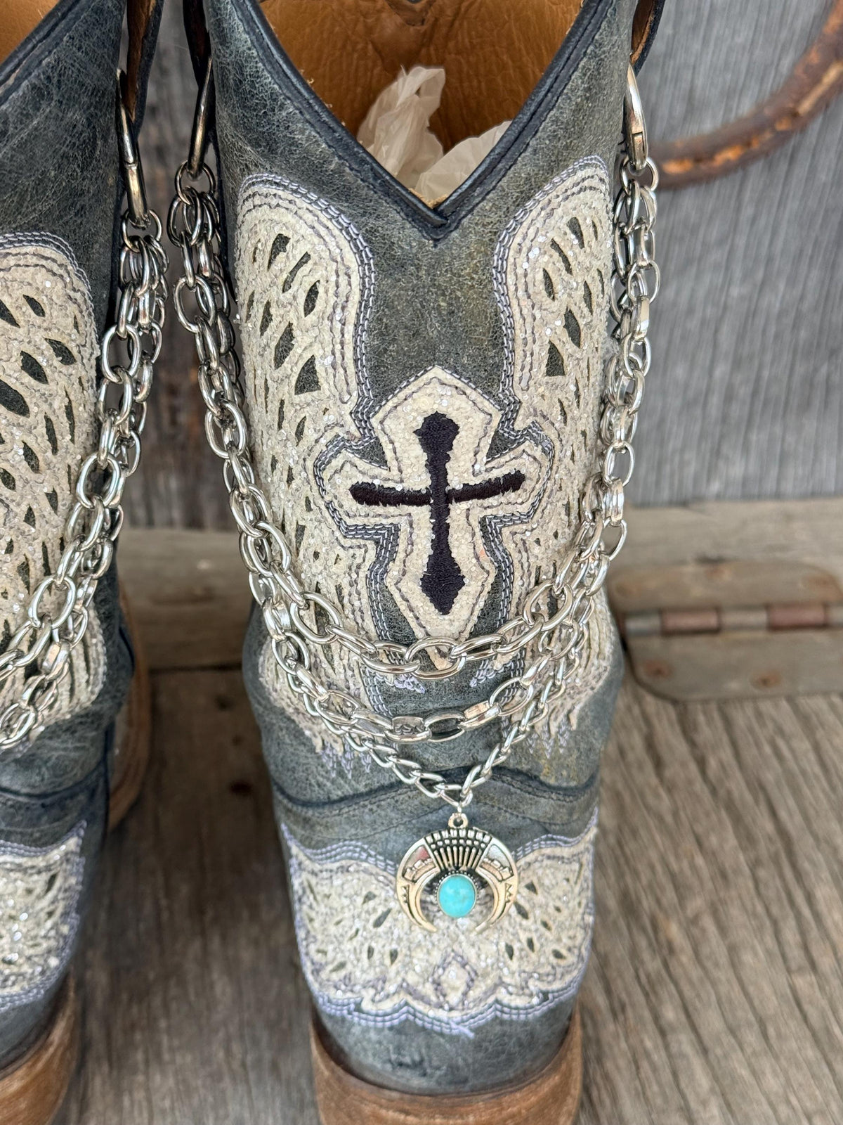 Front-on view of distressed gray cross embroidered cowboy boot with layered silver multi-link chain boot bracelet and turquoise squash blossom charm