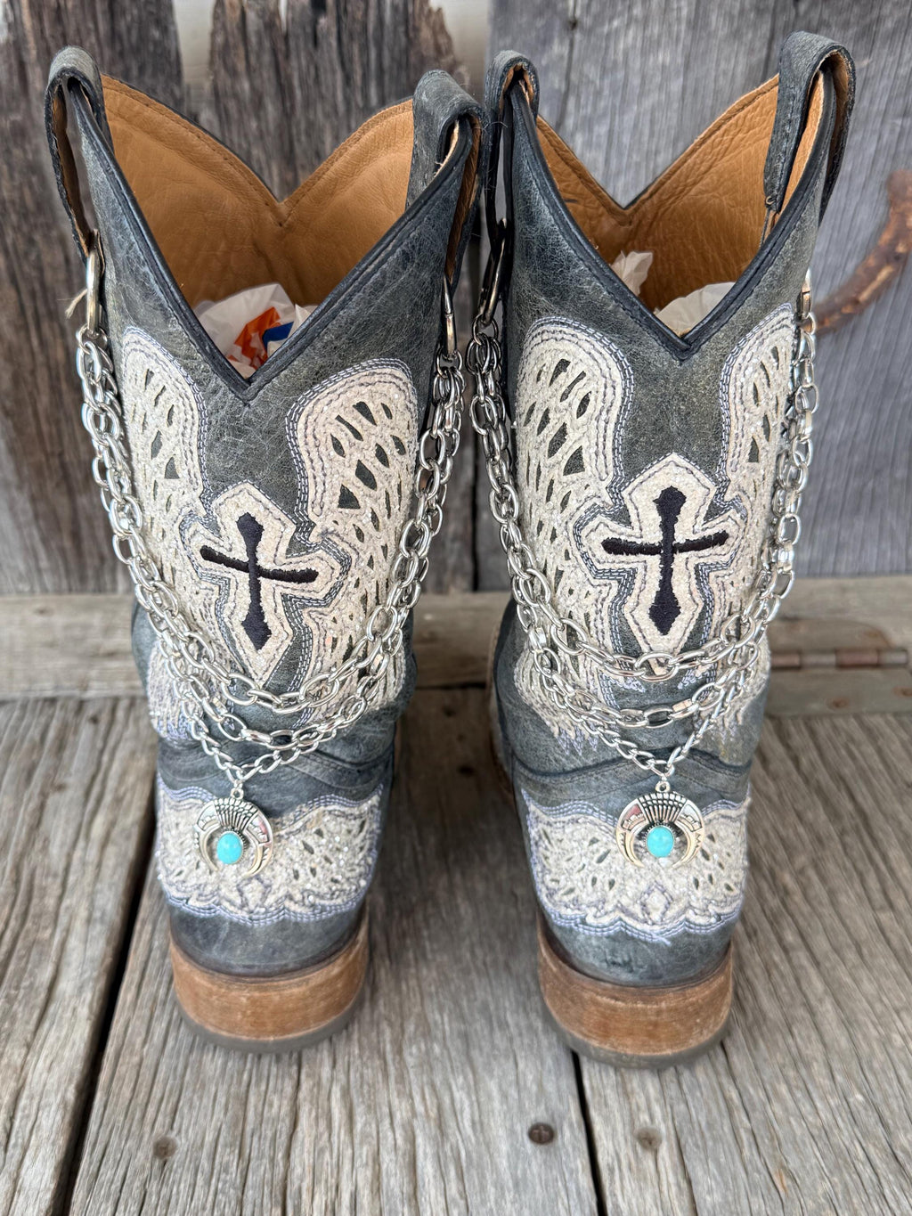 Front view of pair of distressed gray cross and wing embroidered cowboy boots wearing layered silver multi-link chain boot bracelets with turquoise squash blossom charms