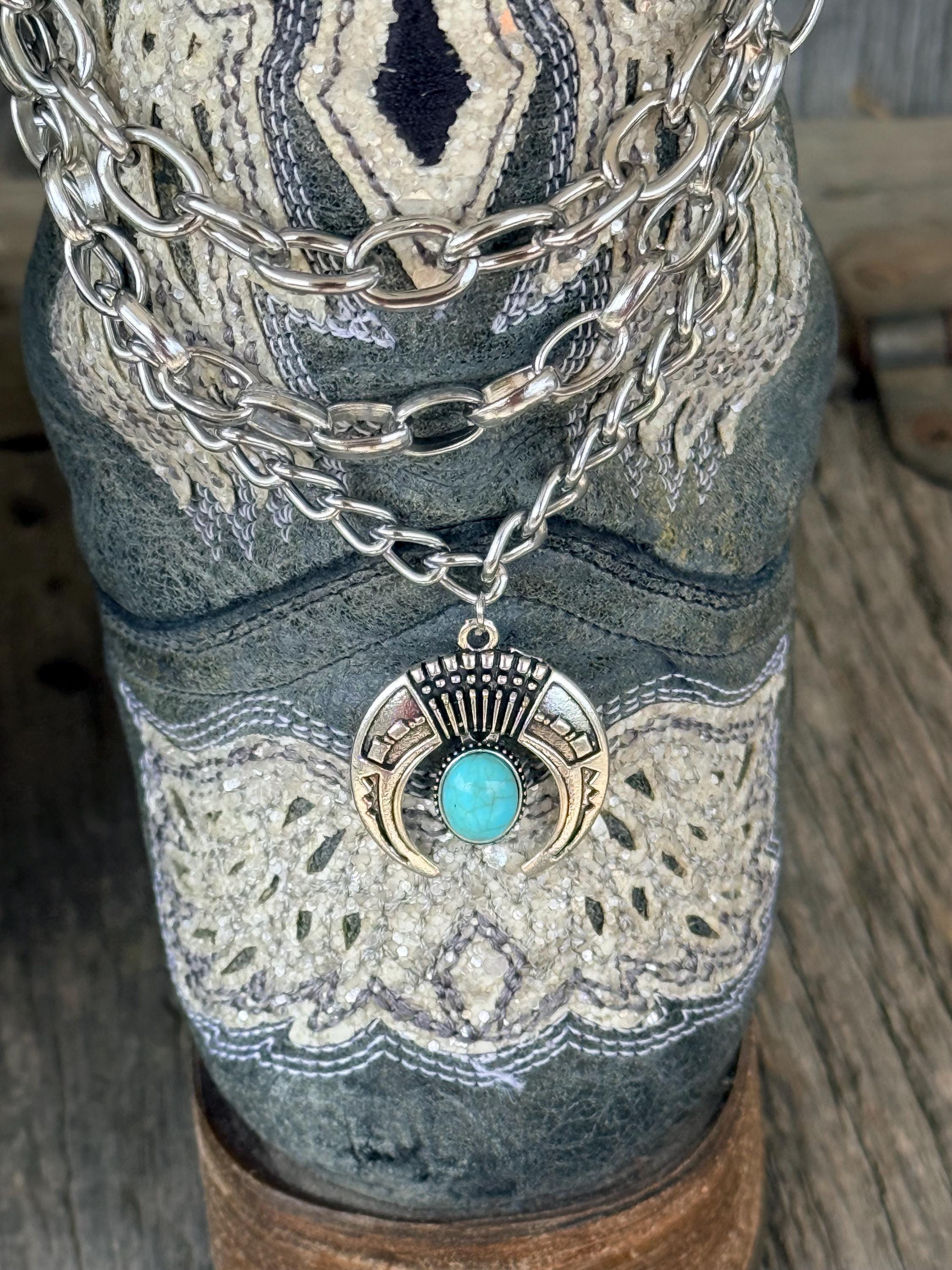 Macro close-up of silver squash blossom charm with turquoise stone center hanging from layered silver chain boot bracelet on distressed blue cowboy boot