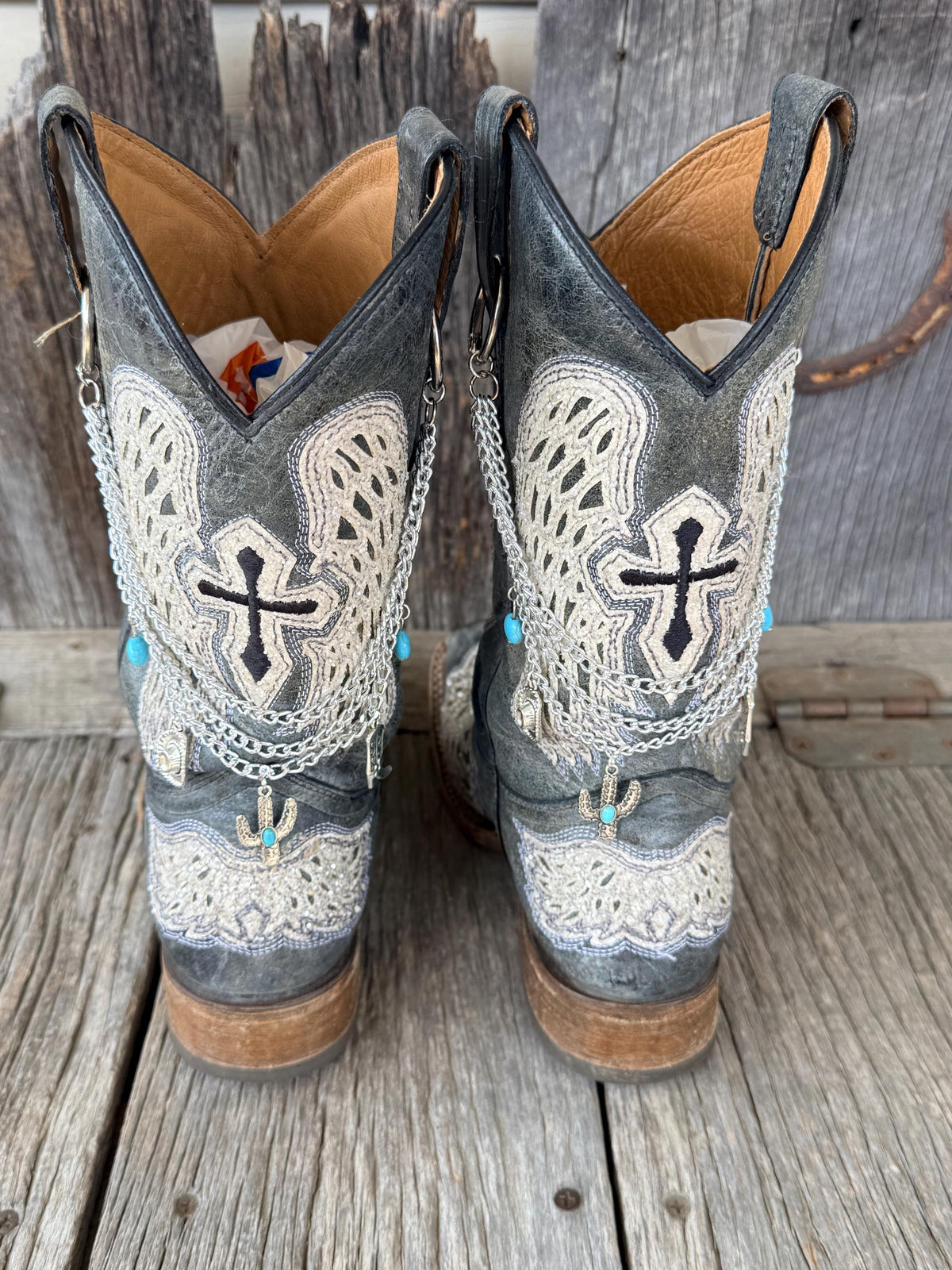 Front view of pair of dark gray distressed cross-embroidered lace-overlay cowboy boots on weathered wood planks wearing three-strand silver chain lei boot bracelets with turquoise oval bead accents and silver cactus charm with turquoise stone, Western boho boot jewelry set of 2