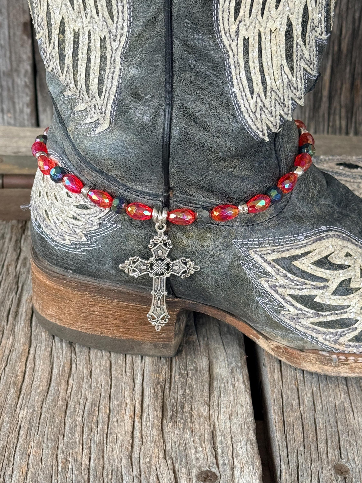 Close-up of dark gray distressed angel wing cowboy boot ankle with red AB faceted oval crystal and dark teal iridescent bead boot bracelet featuring silver rondelle spacers and large ornate silver filigree cross pendant