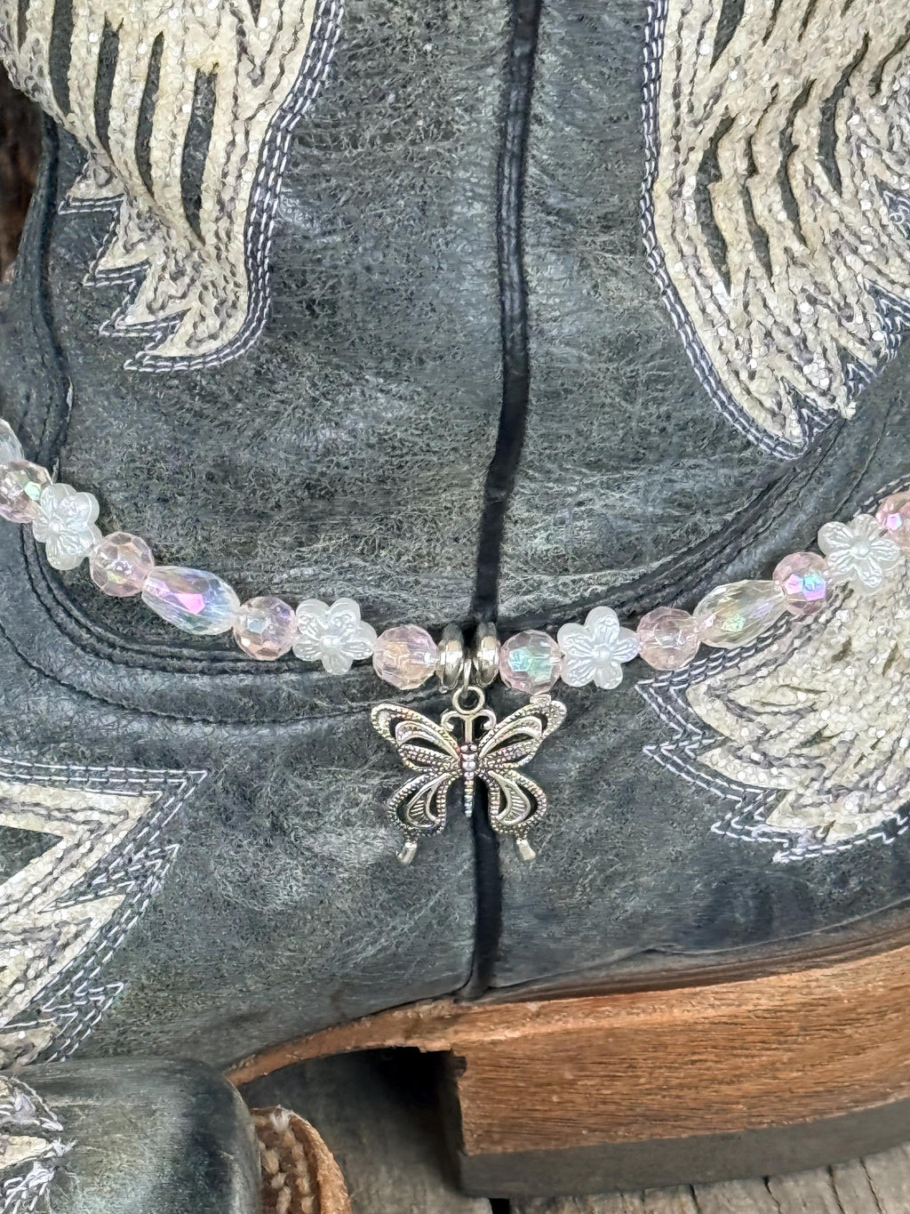Close-up of dark gray distressed angel wing cowboy boot shaft with clear iridescent AB crystal and white flower bead boot bracelet featuring silver filigree butterfly charm pendant, delicate Western boot bling