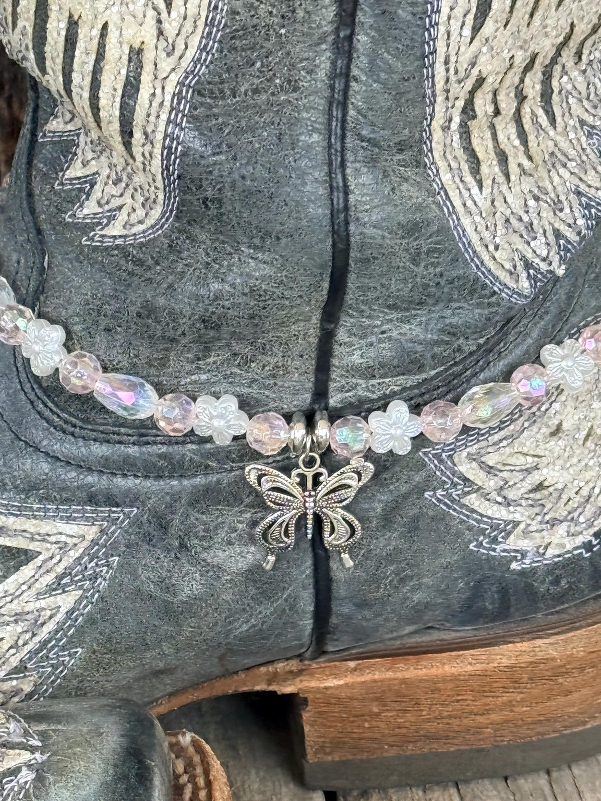 Close-up of dark gray distressed angel wing cowboy boot shaft with clear iridescent AB crystal and white flower bead boot bracelet featuring silver filigree butterfly charm pendant, delicate Western boot bling