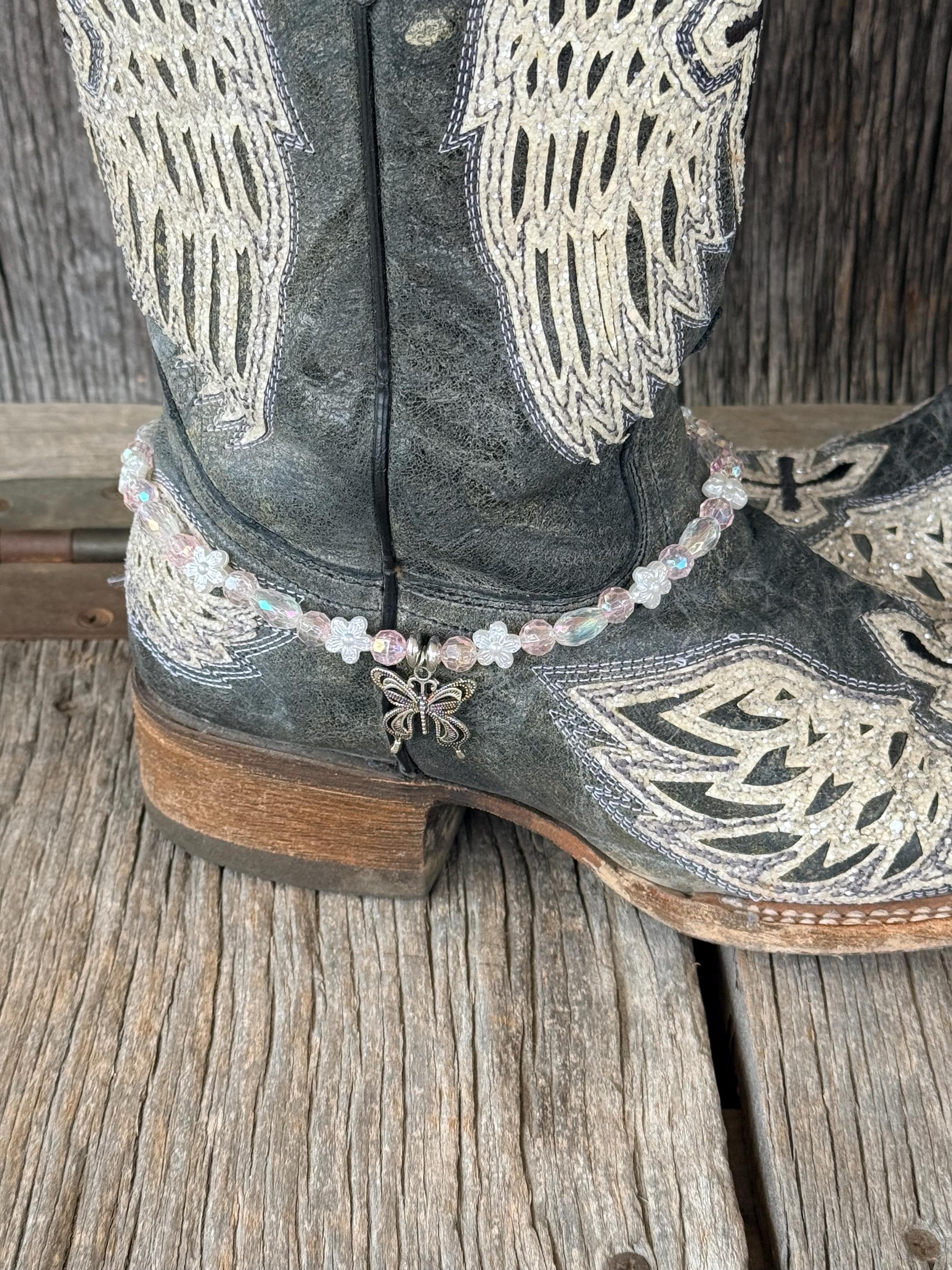 Close-up of dark gray distressed angel wing cowboy boot ankle with clear iridescent AB crystal and white flower bead boot bracelet featuring single silver filigree butterfly charm pendant at center