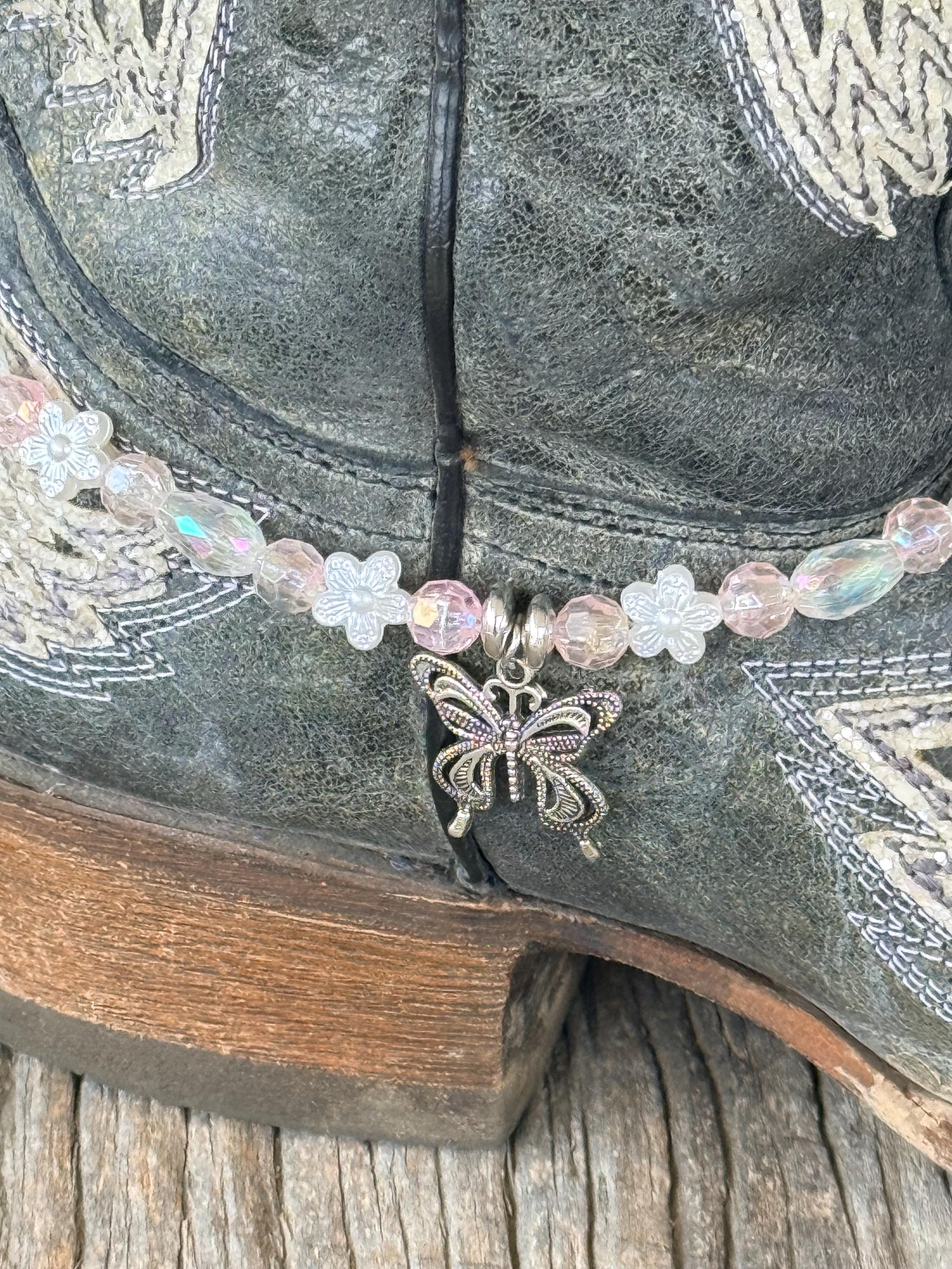 Macro close-up of clear iridescent AB crystal and white flower bead boot bracelet with single silver filigree butterfly charm pendant and silver connector ring, on dark gray distressed angel wing cowboy boot