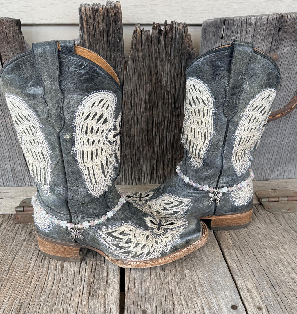 Front view of pair of dark gray distressed angel wing embroidered cowboy boots on weathered wood planks wearing clear iridescent crystal and white flower bead boot bracelets with silver filigree butterfly charm pendant, Western boot bling set of 2