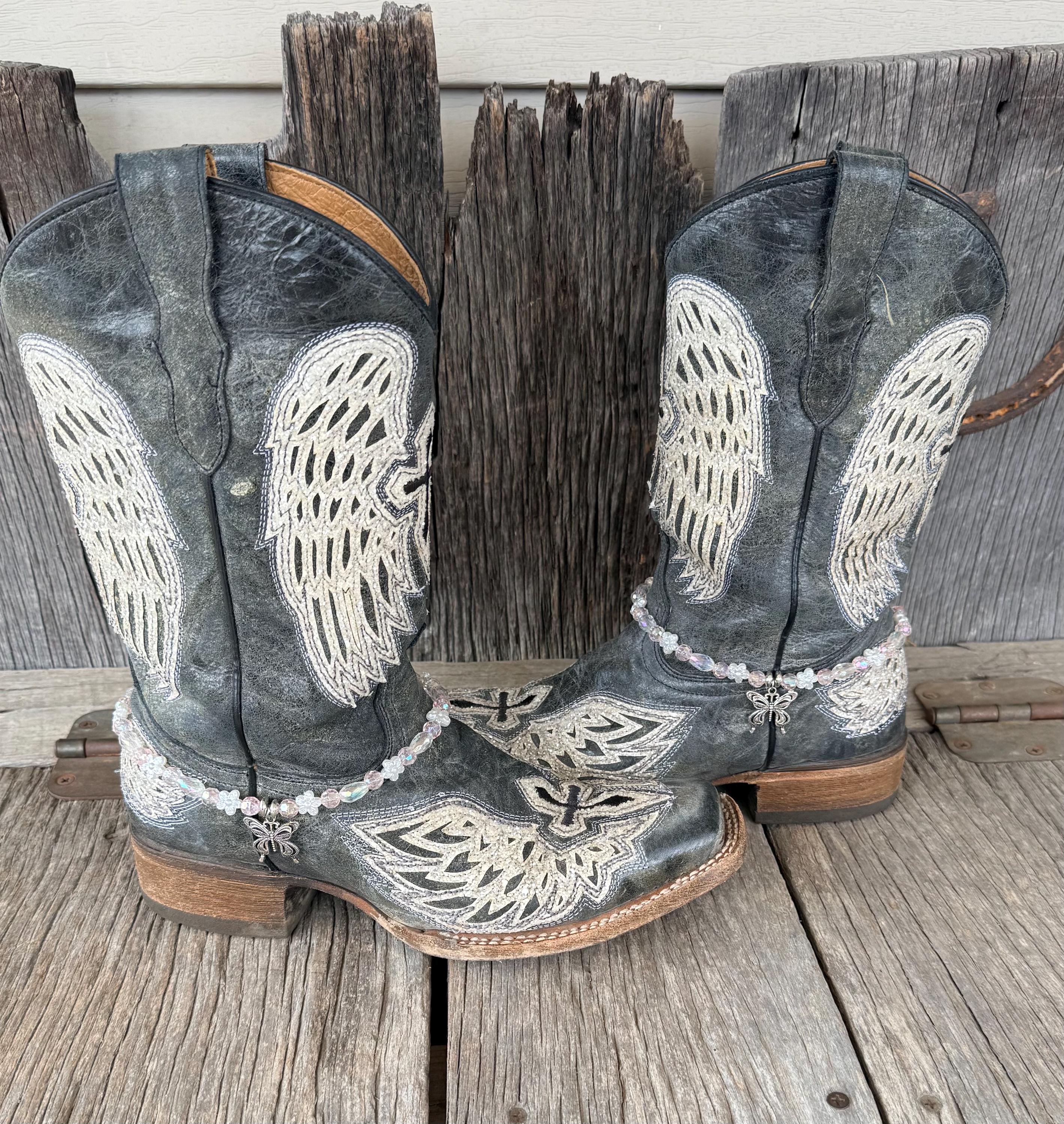 Front view of pair of dark gray distressed angel wing embroidered cowboy boots on weathered wood planks wearing clear iridescent crystal and white flower bead boot bracelets with silver filigree butterfly charm pendant, Western boot bling set of 2