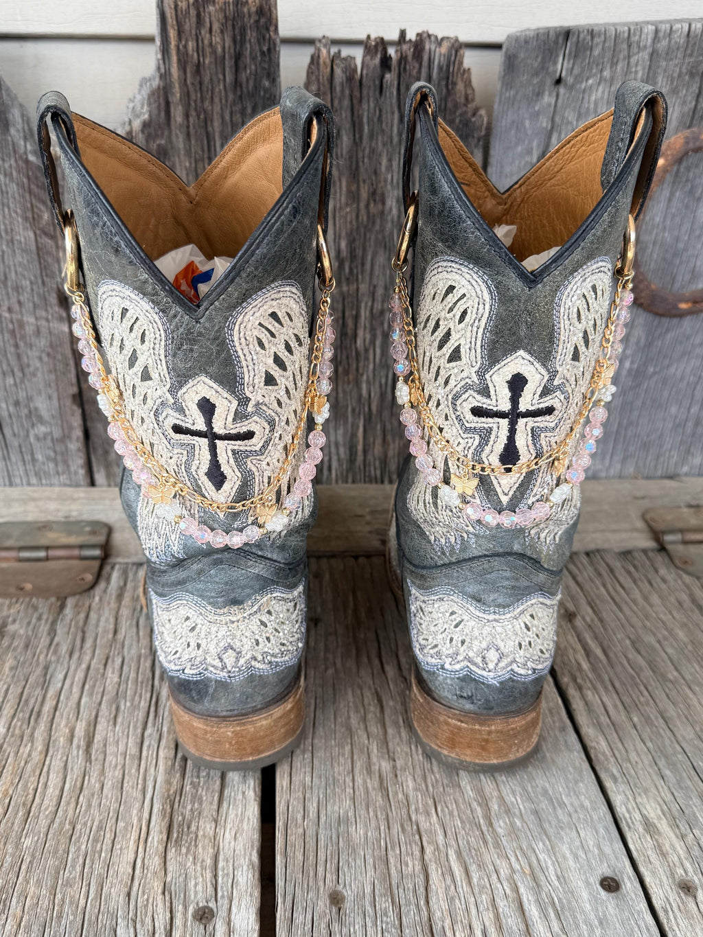 Back view of pair of grey cross embroidered cowboy boots wearing gold link chain and iridescent pink crystal bead boot bracelets with gold butterfly charm, set of 2
