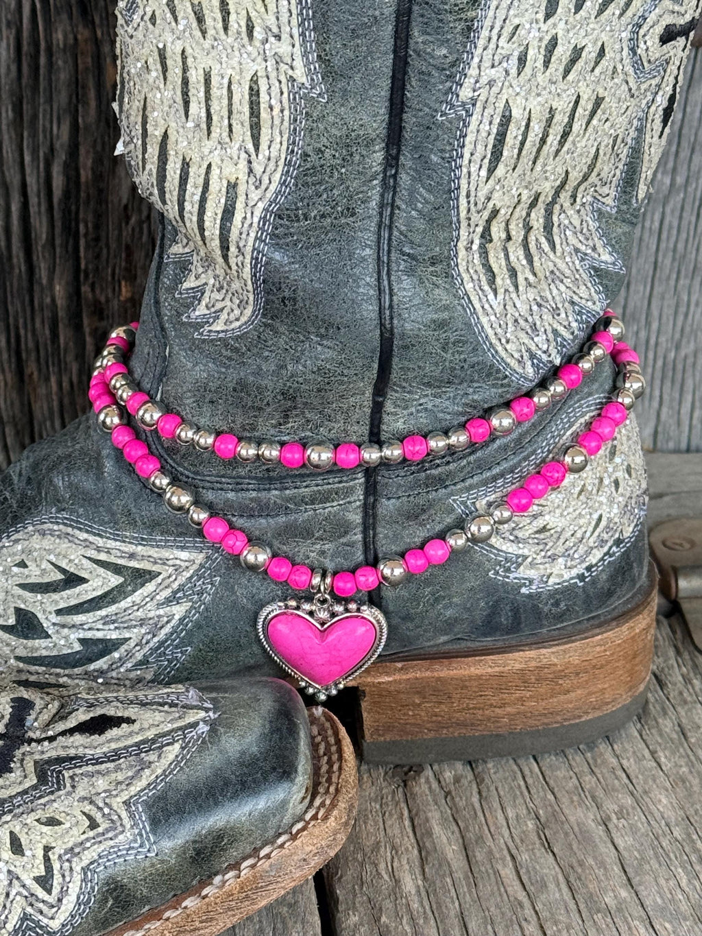 Front view of black distressed angel wing cowboy boot with two-strand hot pink round bead and silver ball spacer boot bracelet chain and large hot pink stone heart pendant in decorative silver rope-edge bezel frame, Western boot bling