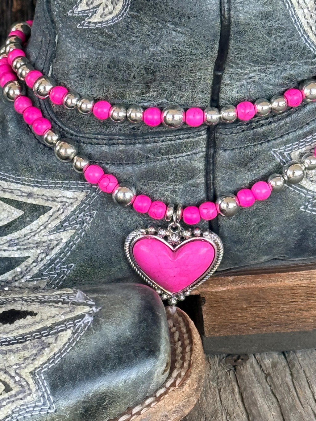 Macro close-up of two-strand hot pink round bead and silver ball spacer boot bracelet chain with large hot pink stone heart pendant set in decorative silver rope-edge bezel frame, on black distressed cowboy boot