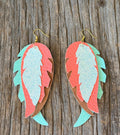 A pair of handmade feather-shaped drop earrings made from layered textured leather in coral pink, mint green, and brown, hanging from gold fishhook wires on a rustic weathered wood background.