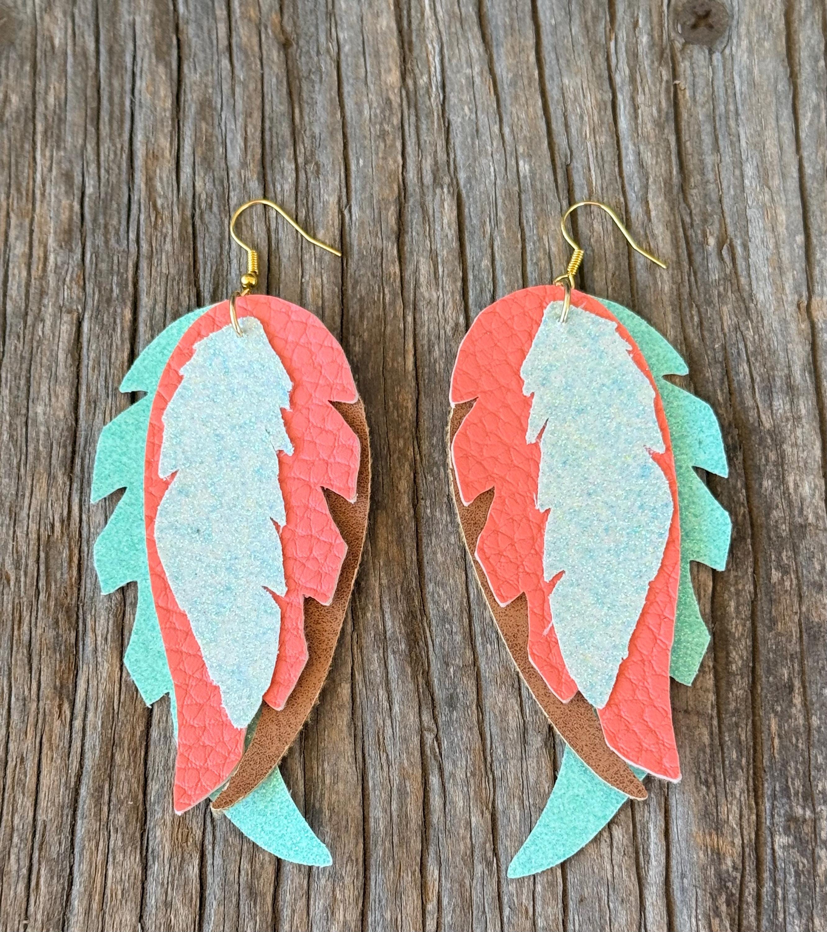 A pair of handmade feather-shaped drop earrings made from layered textured leather in coral pink, mint green, and brown, hanging from gold fishhook wires on a rustic weathered wood background.