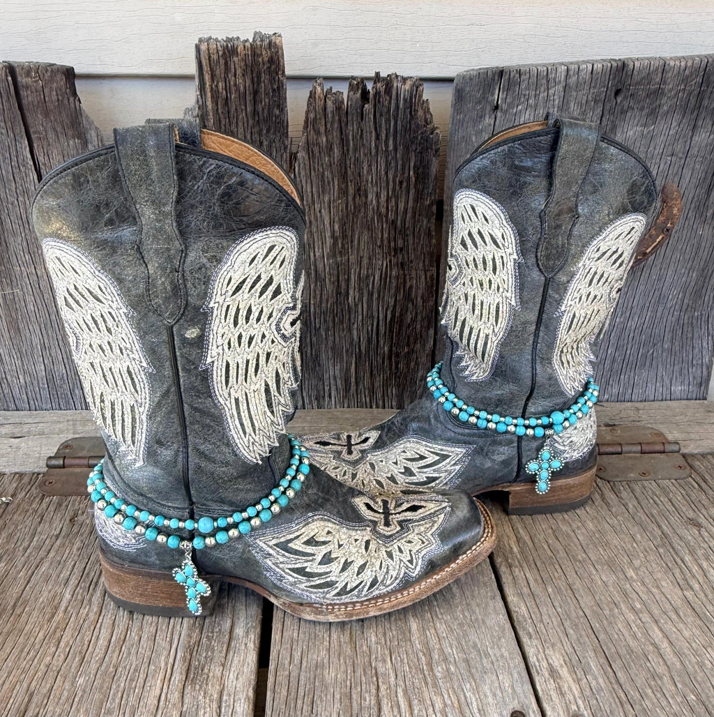 Pair of black angel wing embroidered cowboy boots wearing silver and turquoise beaded boot bracelets with turquoise cross charms, set of 2