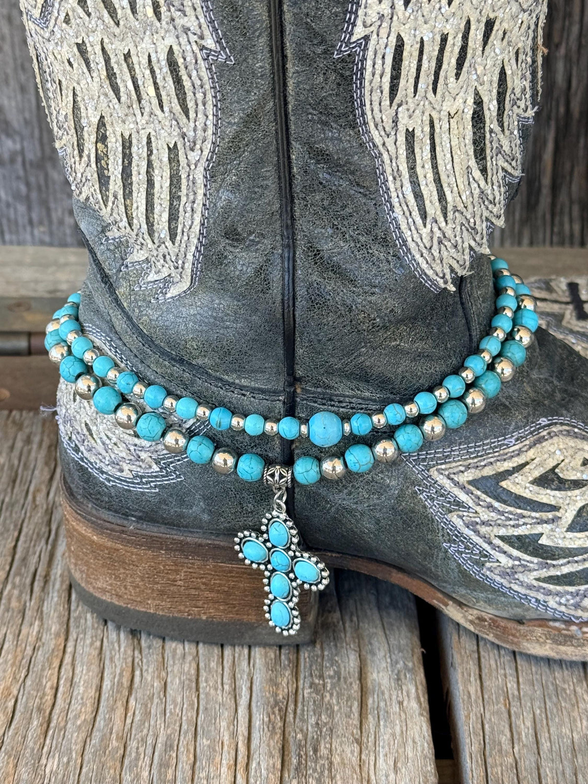 Close-up of double-strand silver and turquoise beaded boot bracelet on black angel wing cowboy boot with turquoise stone cross charm pendant