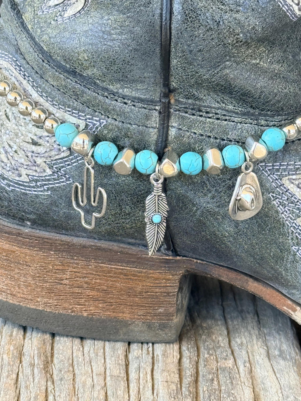 Macro close-up of turquoise stone and silver bead boot bracelet with three silver Western charms: cactus outline, feather with turquoise bead accent, and cowboy hat silhouette, on black distressed cowboy boot
