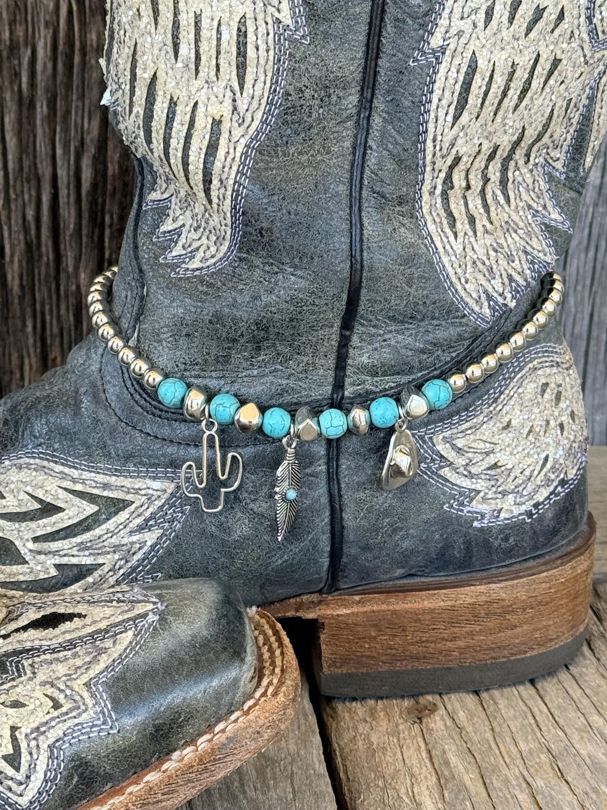 Side view of black distressed angel wing embroidered cowboy boot on wood planks with turquoise stone and silver bead boot bracelet featuring silver cactus, feather with turquoise accent, and cowboy hat charms