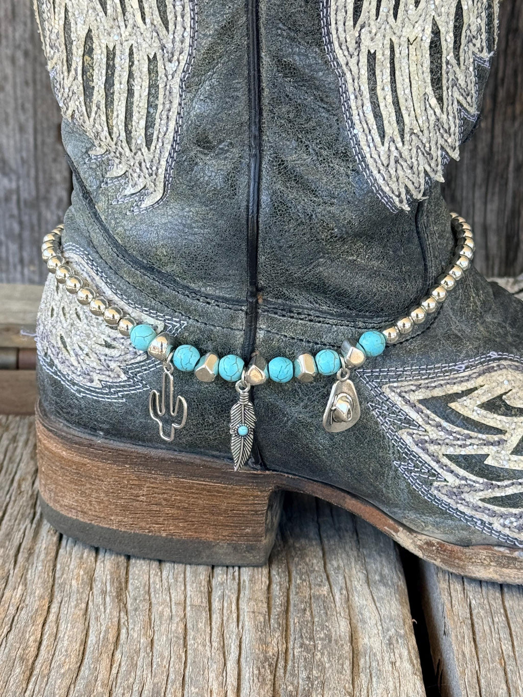 Close-up of black distressed angel wing cowboy boot ankle with turquoise stone and silver bead boot bracelet featuring three silver charms: cactus, feather with turquoise accent, and cowboy hat