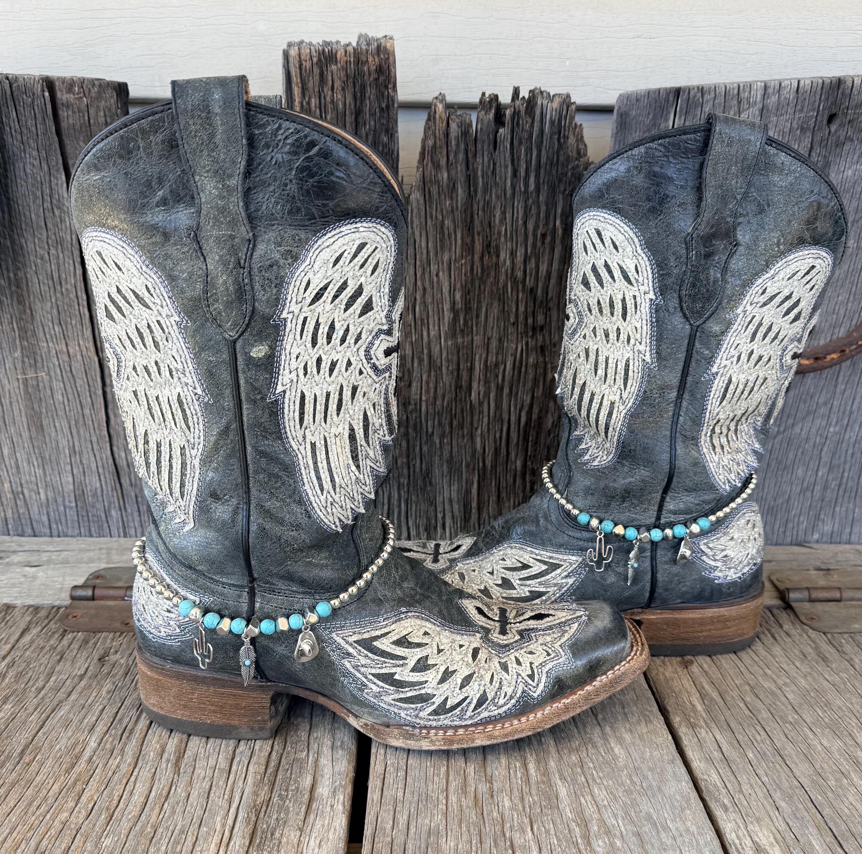 Front view of pair of black distressed angel wing embroidered cowboy boots on rustic wood planks wearing turquoise stone and silver bead boot bracelets with silver cactus, feather, and cowboy hat charms, set of 2