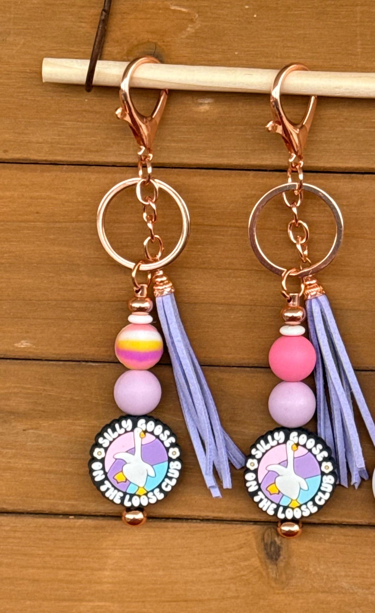 A pair of dangling keychains with rose gold-tone hardware, lavender tassels, pastel beads, and circular swan-themed charms, displayed on a wooden surface. Silly Goose On The Loose Club.