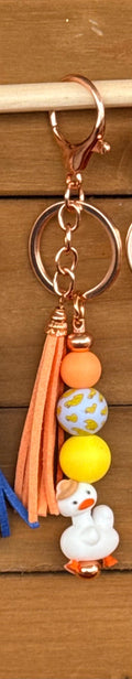 A close-up of a keychain with a rose gold clasp and key ring, featuring a peach tassel, orange and yellow beads, a patterned blue and white duck bead, and a white goose charm on a wooden background.