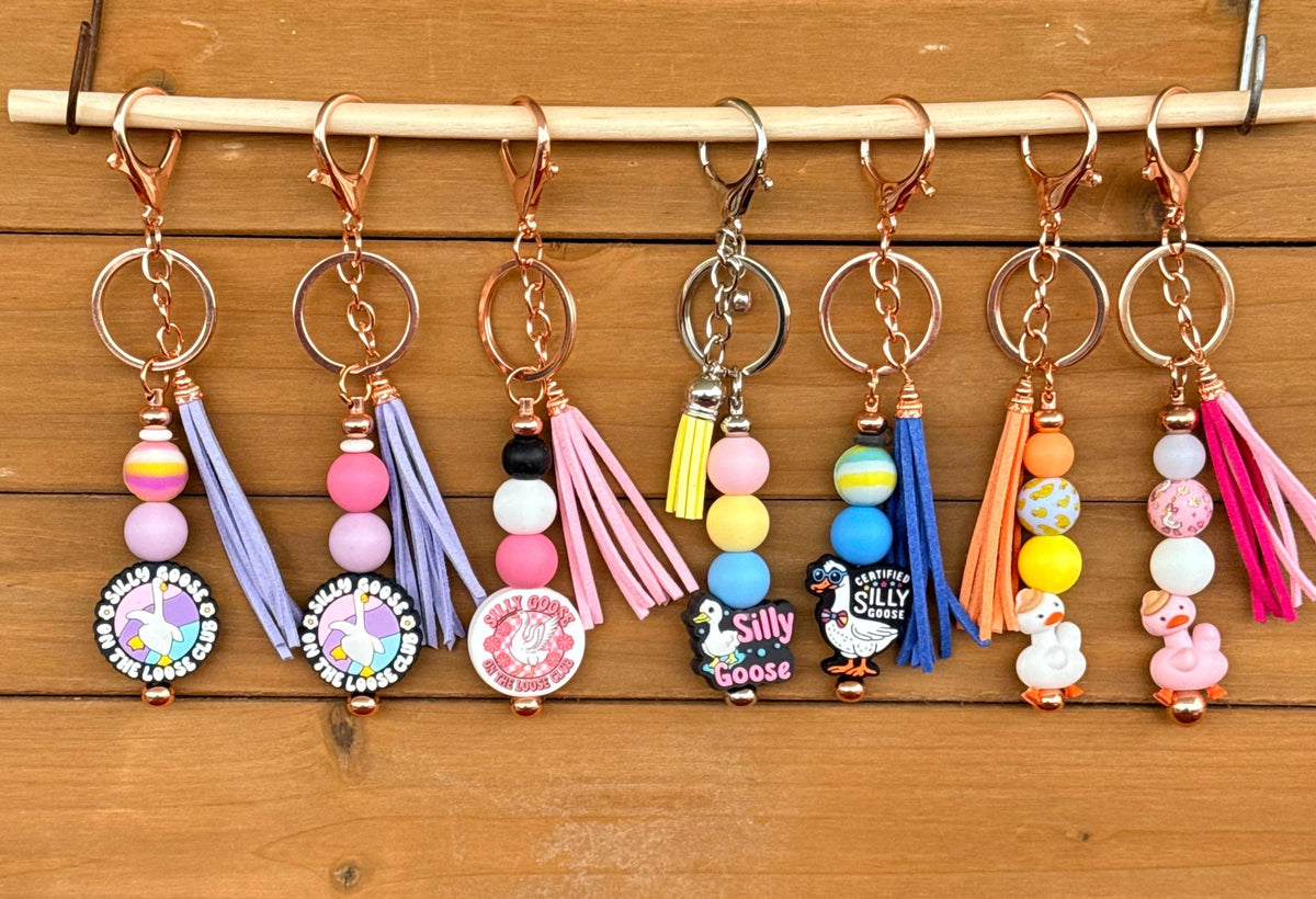 Seven colorful beaded keychains with tassels and round charms hanging from a wooden dowel on a wood plank background.