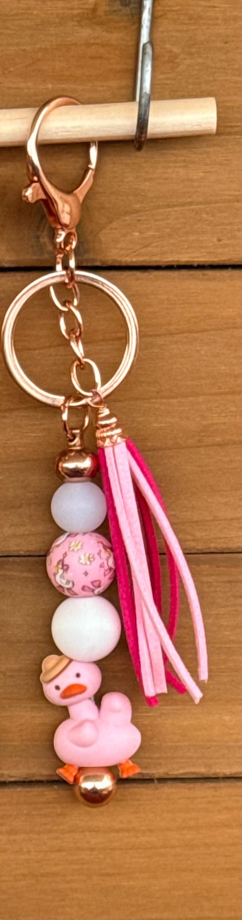 A pink beaded keychain with a copper-toned clasp, a split key ring, a tassel, and a small goose charm, shown against a wooden background.