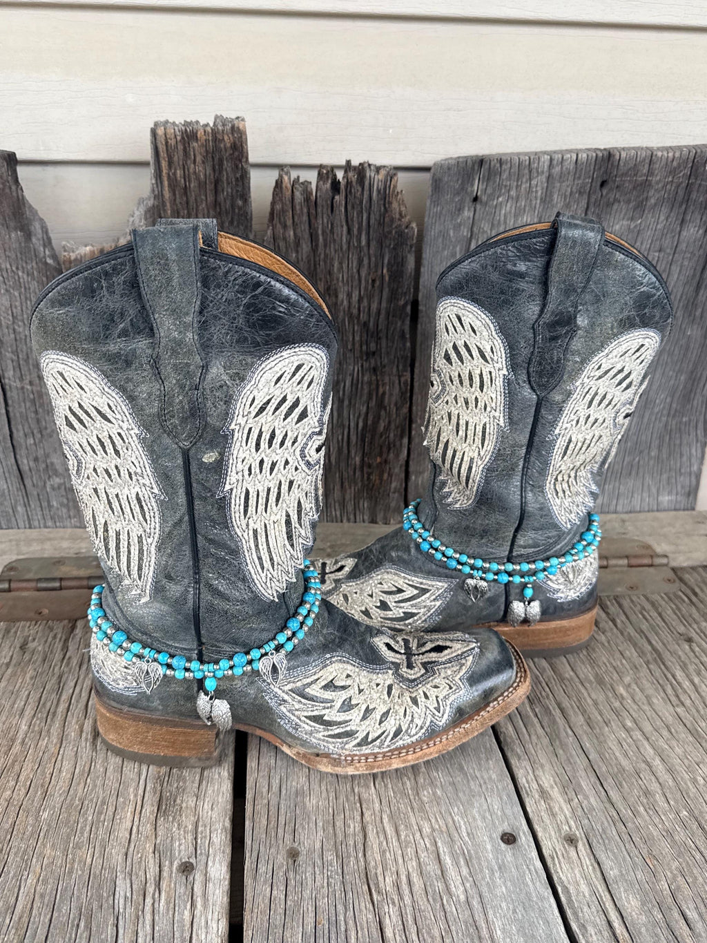 A pair of distressed gray western cowboy boots with white embroidered wing and floral patterns, decorated with turquoise beaded ankle bracelets and silver angel wing charms, set against a rustic wooden background.