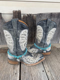 A pair of distressed gray western cowboy boots with white embroidered wing and floral patterns, decorated with turquoise beaded ankle bracelets and silver angel wing charms, set against a rustic wooden background.