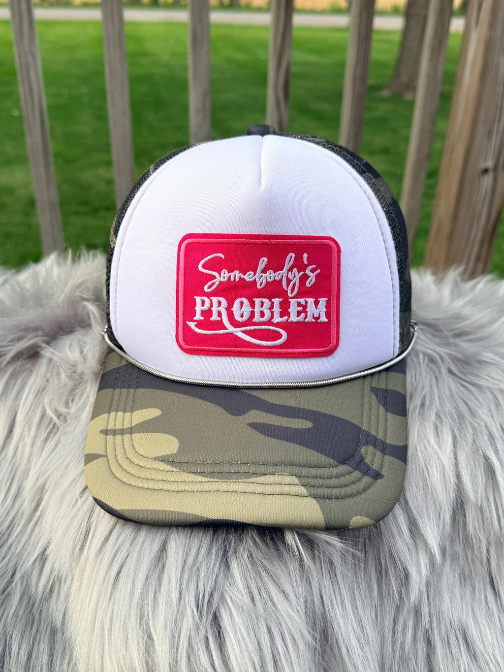 A white and black trucker hat with a camouflage brim, featuring a red square patch on the front with white script and block lettering. 